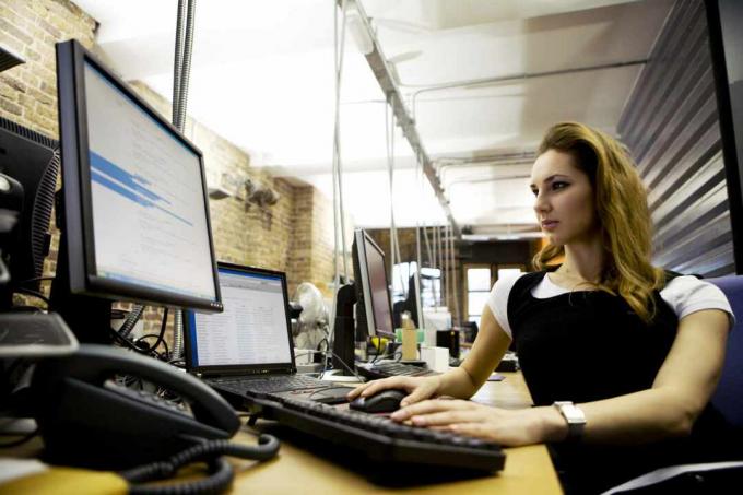 Una mujer sentada en un escritorio usando una computadora portátil con teclado externo y monitor.