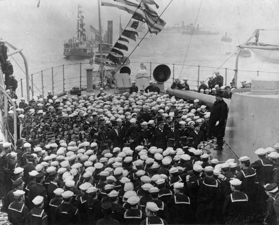 El presidente Theodore Roosevelt parado en una torreta de acorazado con una multitud de marineros frente a él.